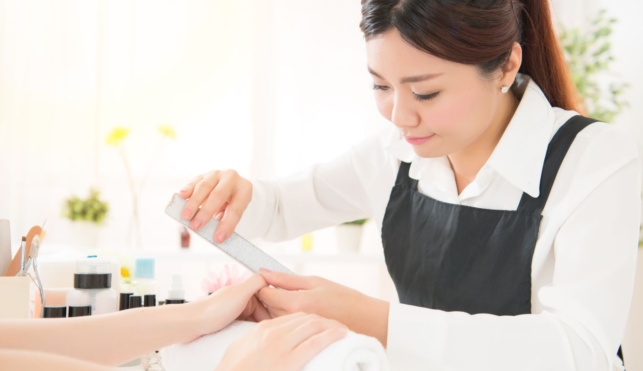 Photo of a young nail technician filing a customer's fingernails in a salon