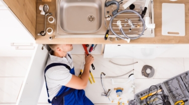 Photo of a young plumber examining the underside of a kitchen sink with some tools