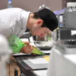 Culinary arts competitor with white uniform and black cap piping mix onto a baking tray using a green piping bag in a commercial kitchen, with baking equipment and mixers in the background.