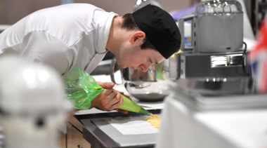 Culinary arts competitor with white uniform and black cap piping mix onto a baking tray using a green piping bag in a commercial kitchen, with baking equipment and mixers in the background.
