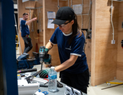 Female competitor wearing safety glasses drills a metal piece at a workbench in a workshop, with electrical training walls in the background.