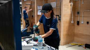 Female competitor wearing safety glasses drills a metal piece at a workbench in a workshop, with electrical training walls in the background.