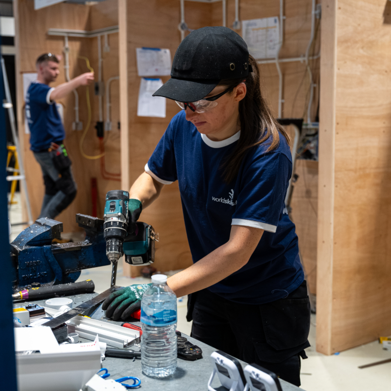 Female competitor wearing safety glasses drills a metal piece at a workbench in a workshop, with electrical training walls in the background.