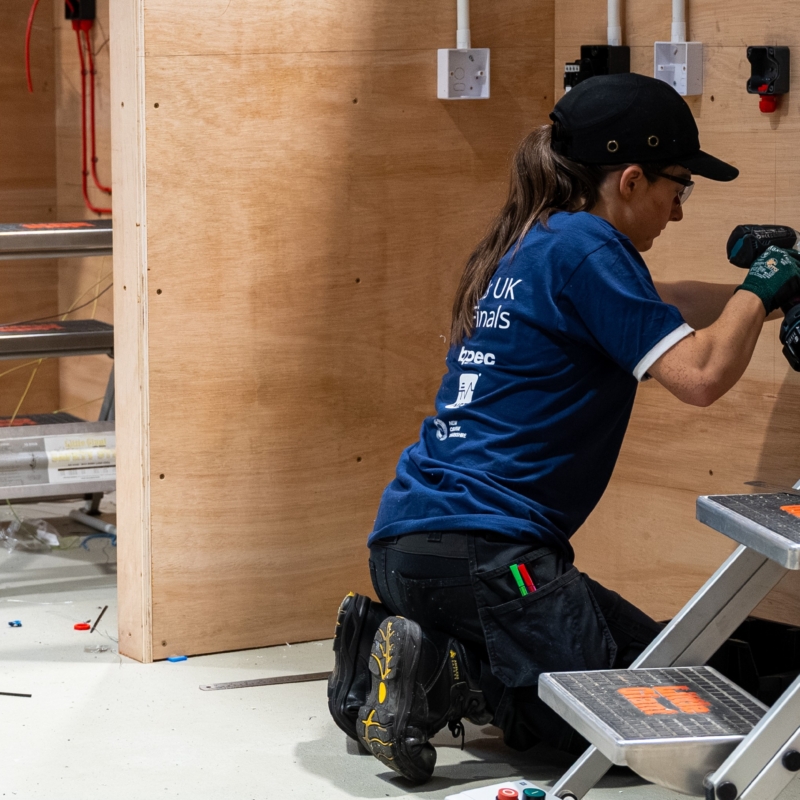 Female competitor using a power drill to install electrical components on a wooden wall in an indoor workshop.