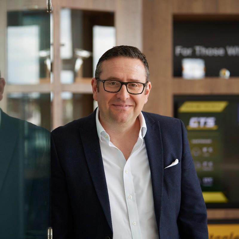 General Manager at StanleyBlack & Decker UK & ROI, wearing glasses, a navy blazer, and a white open-collar shirt smiles at the camera while standing in a contemporary office.