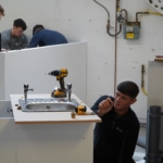 A group of plumbing professionals work together to install and adjust a sink unit in a workshop, with tools and fittings arranged on the worktop.