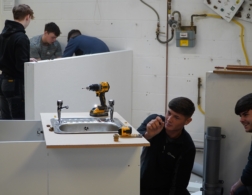 A group of plumbing professionals work together to install and adjust a sink unit in a workshop, with tools and fittings arranged on the worktop.