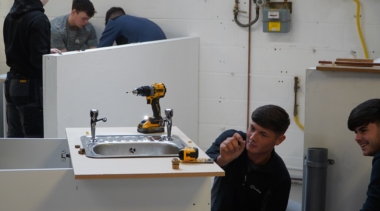 A group of plumbing professionals work together to install and adjust a sink unit in a workshop, with tools and fittings arranged on the worktop.