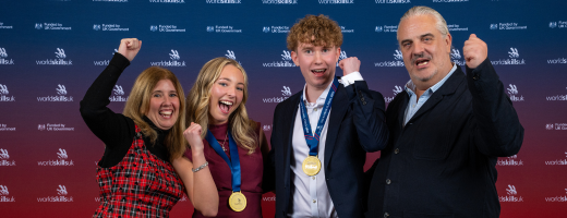 Four people pose in front of a WorldSkills UK event backdrop, smiling and raising their fists in celebration. Two young medalists wearing gold medals stand in the center.