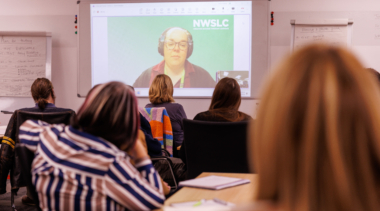 A group of people at a workshop, watching a presenter on a screen during a video call.
