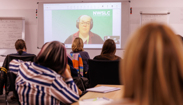 A group of people at a workshop, watching a presenter on a screen during a video call.